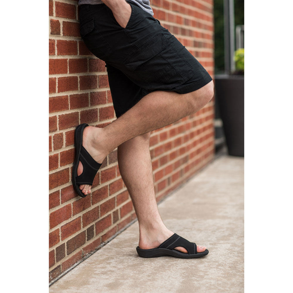 PowerStep Orthotic Arch Supporting Slide Sandals for Men, man standing against brick wall with foot kicked back on wall while wearing black slide sandals with arch support