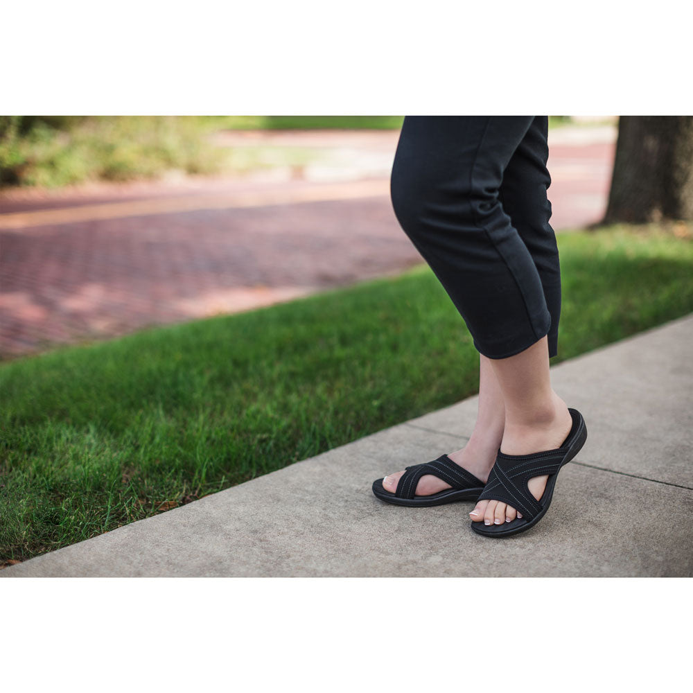 PowerStep Orthotic Arch Supporting Slide Sandals for Women, woman standing on sidewalk in black slide sandals with green grass and brick road in the background